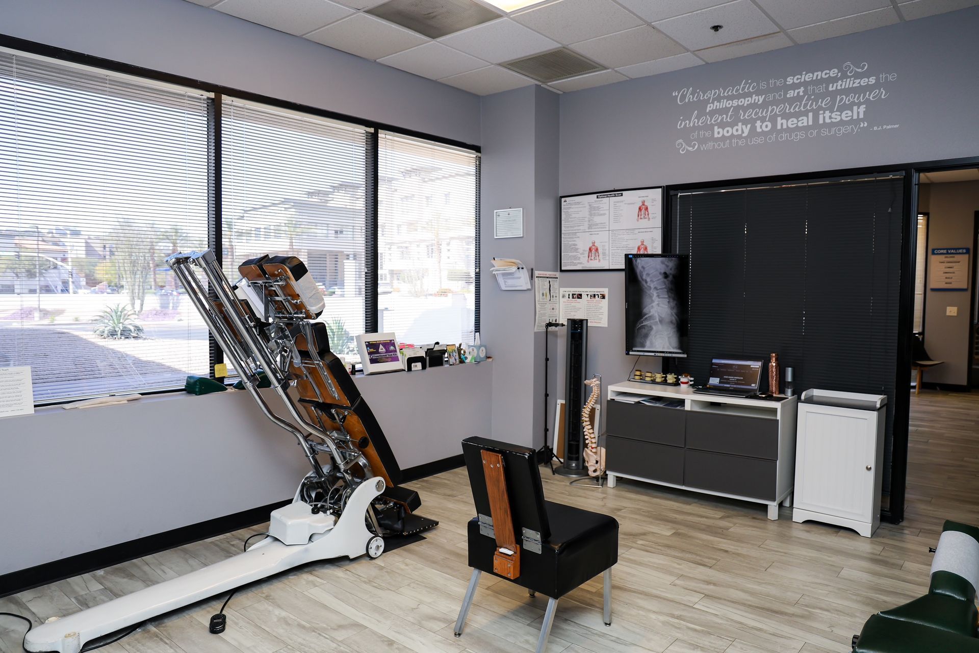 A medical office featuring a physical therapy table, a black chair, medical charts, and anatomical models.