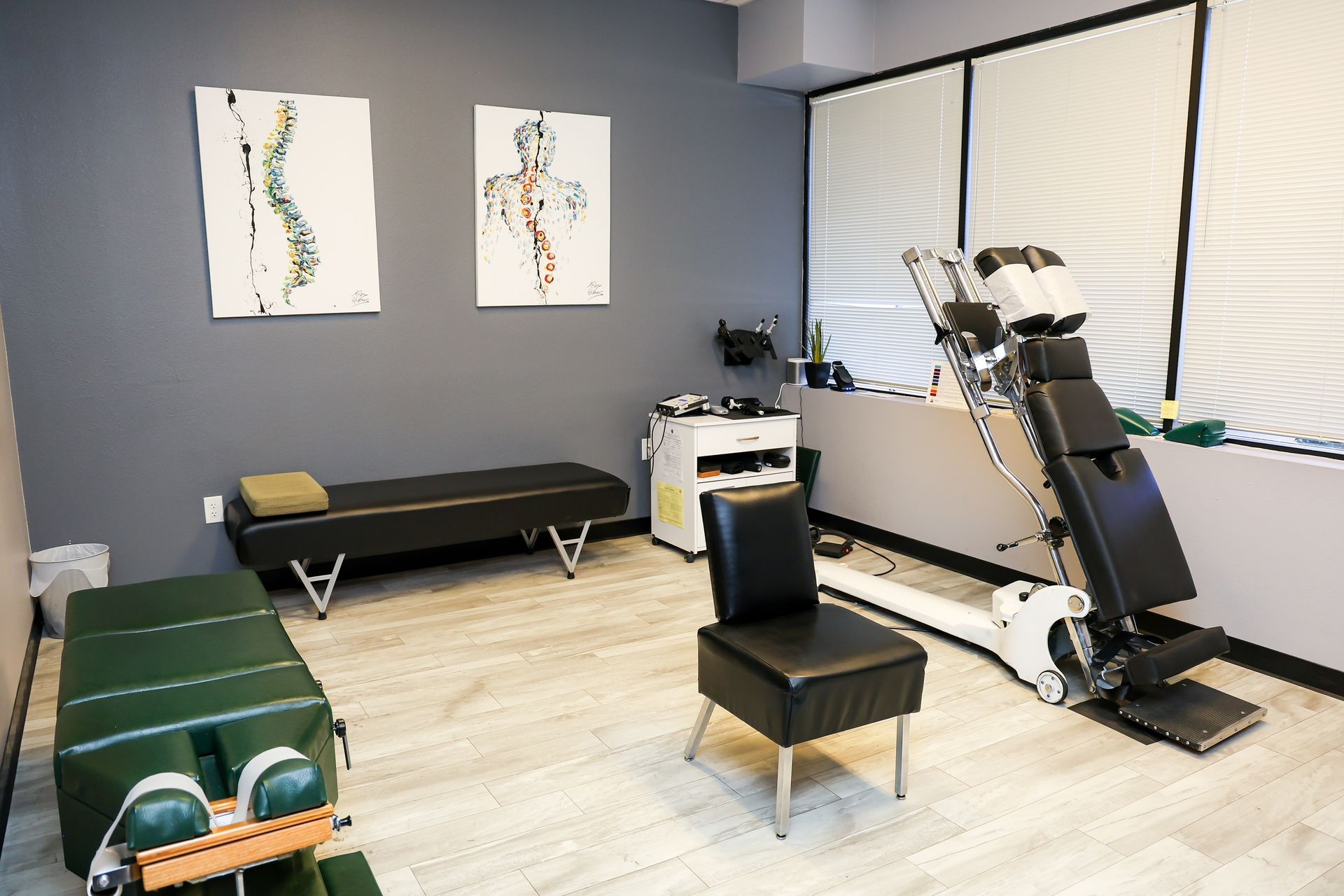 A chiropractic office with a treatment table, specialized spinal adjustment equipment, a chair, and anatomical wall art.
