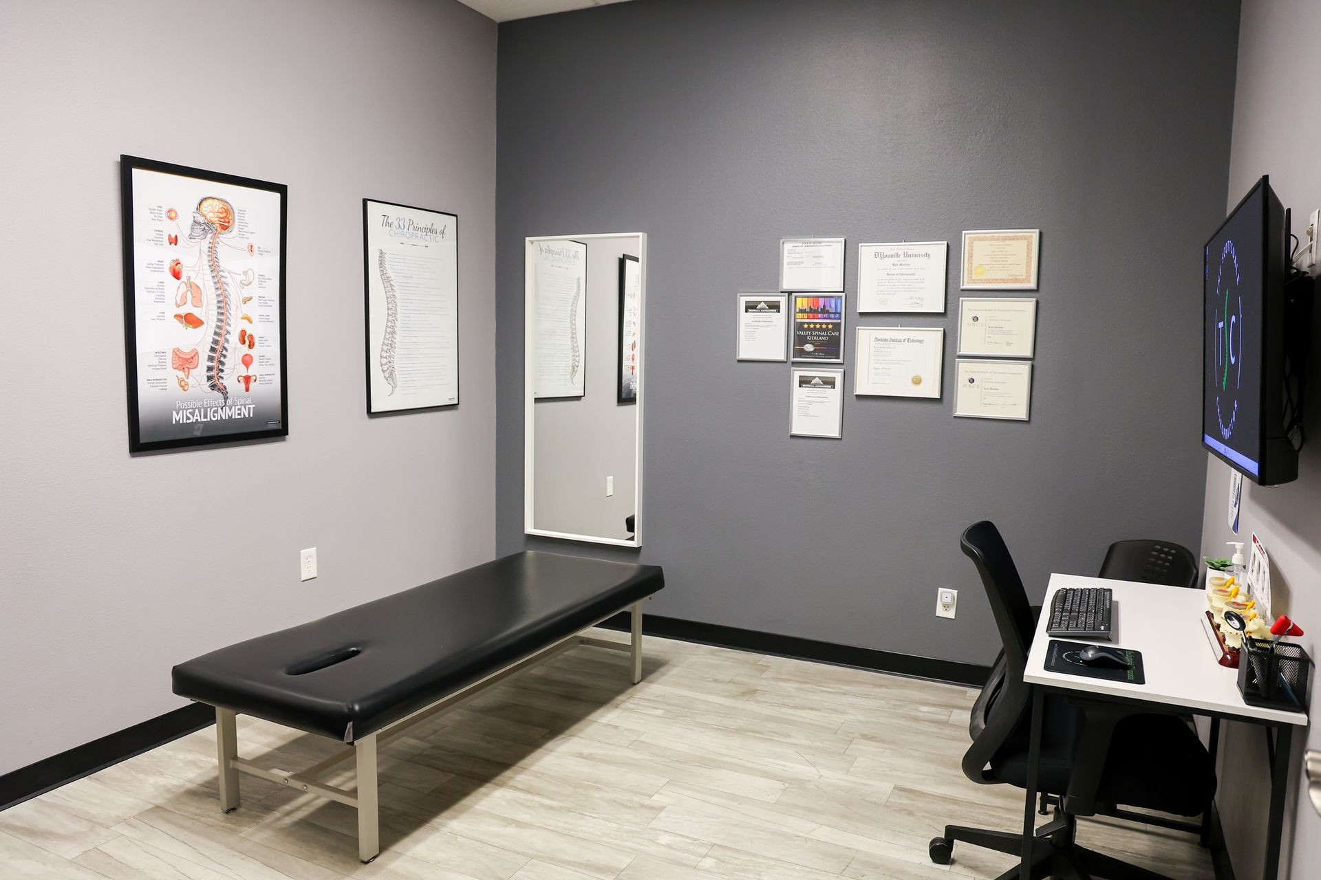 A chiropractic exam room with a black treatment table, wall charts of the spine, framed certifications, and a desk.