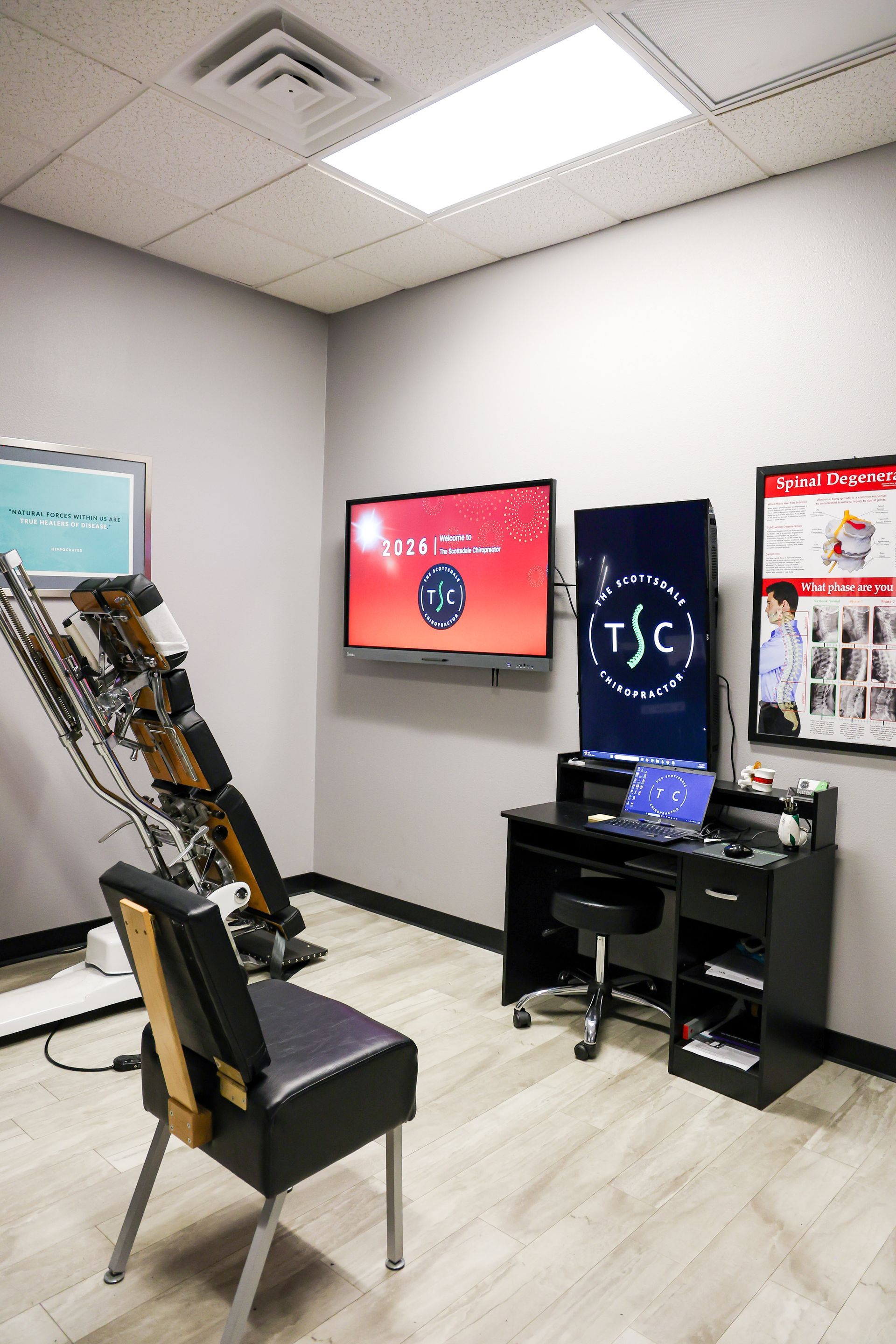 A treatment room with a chiropractic table, a desk with a computer, and medical wall charts under bright lighting.