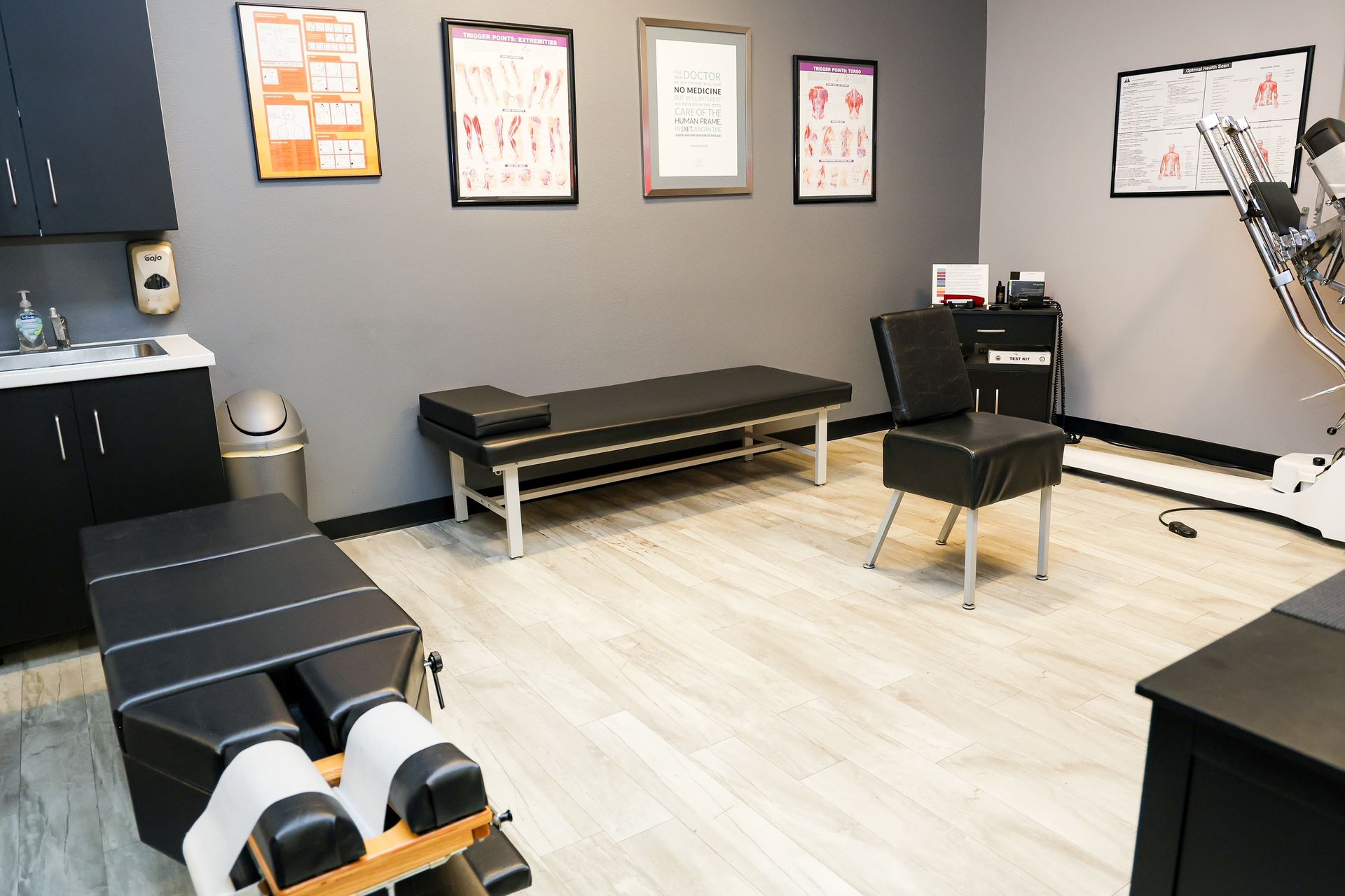 A chiropractic treatment room with black exam tables, a chair, and anatomical posters on gray walls.