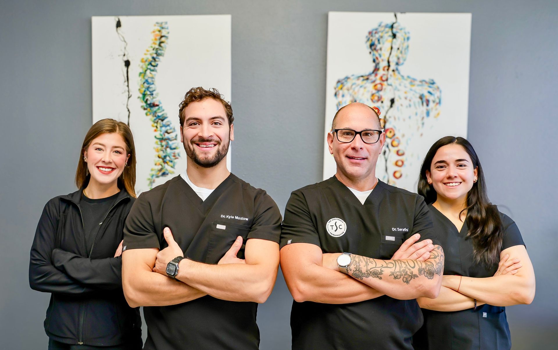 Four professional staff members stand with arms crossed in front of two abstract spinal column art pieces on a gray wall.