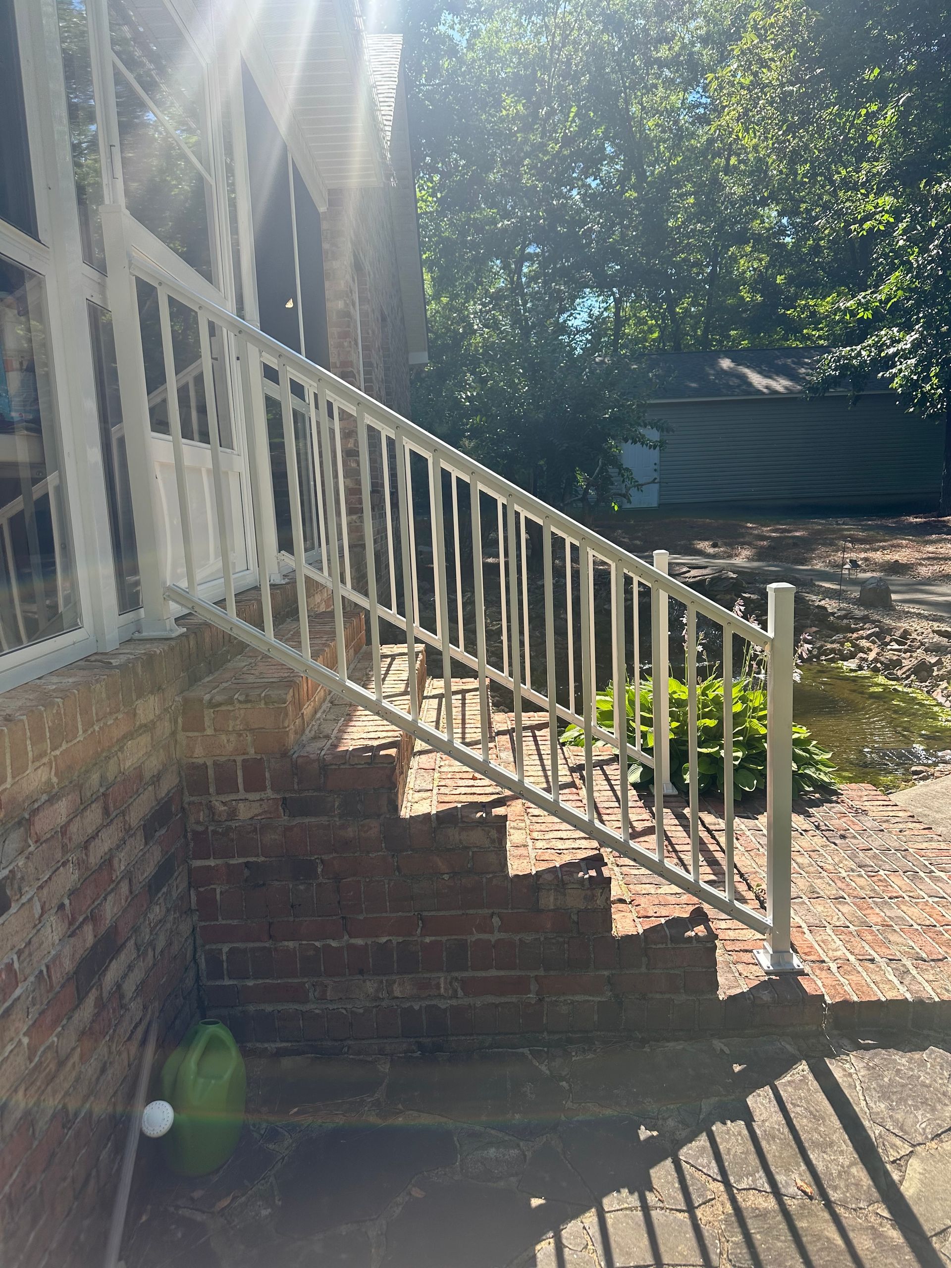 Brick house steps with white railing in sunny yard, with brick wall and trees in the background.