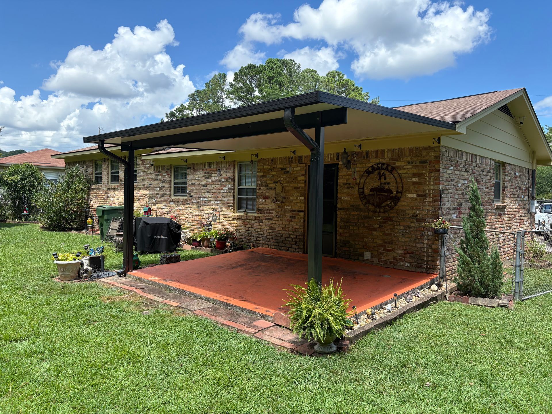 Brick house patio with black metal roof, red floor, and green lawn under a partly cloudy sky