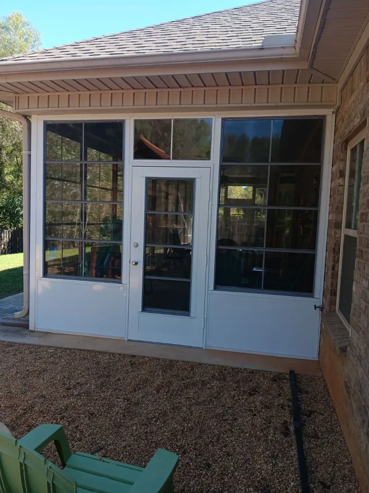 Front porch with white screened door and windows, under a tan roof, bordered by brick and gravel.