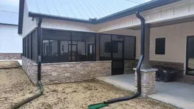 Covered patio with stone half-wall, black-framed windows, and concrete walkway beside a house.