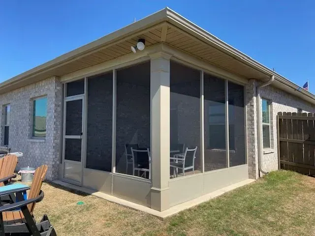 Covered patio attached to a beige brick house, with screened windows and outdoor chairs on a grassy yard.