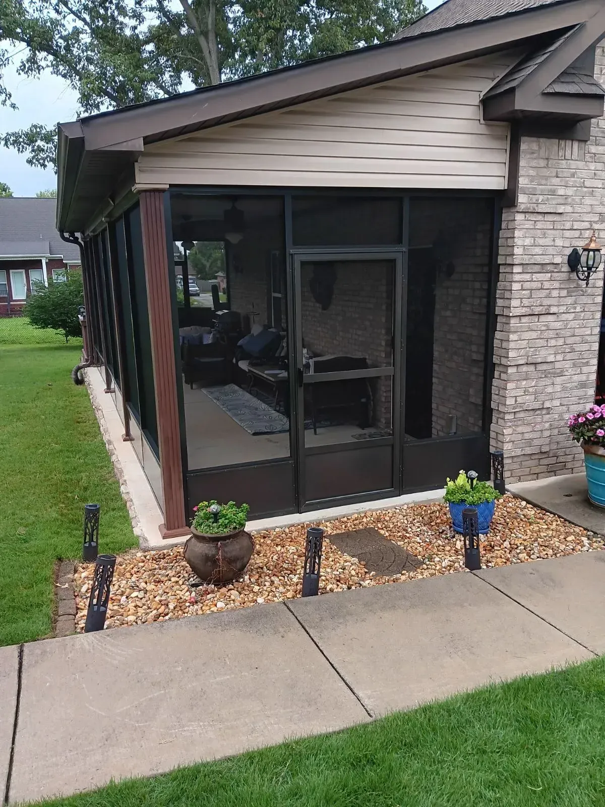 Screened front porch of a house with potted plants and a stone facade beside a sidewalk.