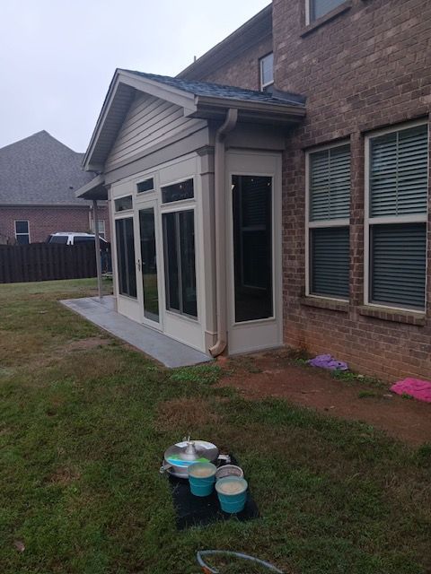 Backyard patio beside a brick house, with windows, grass, and paint buckets in the foreground