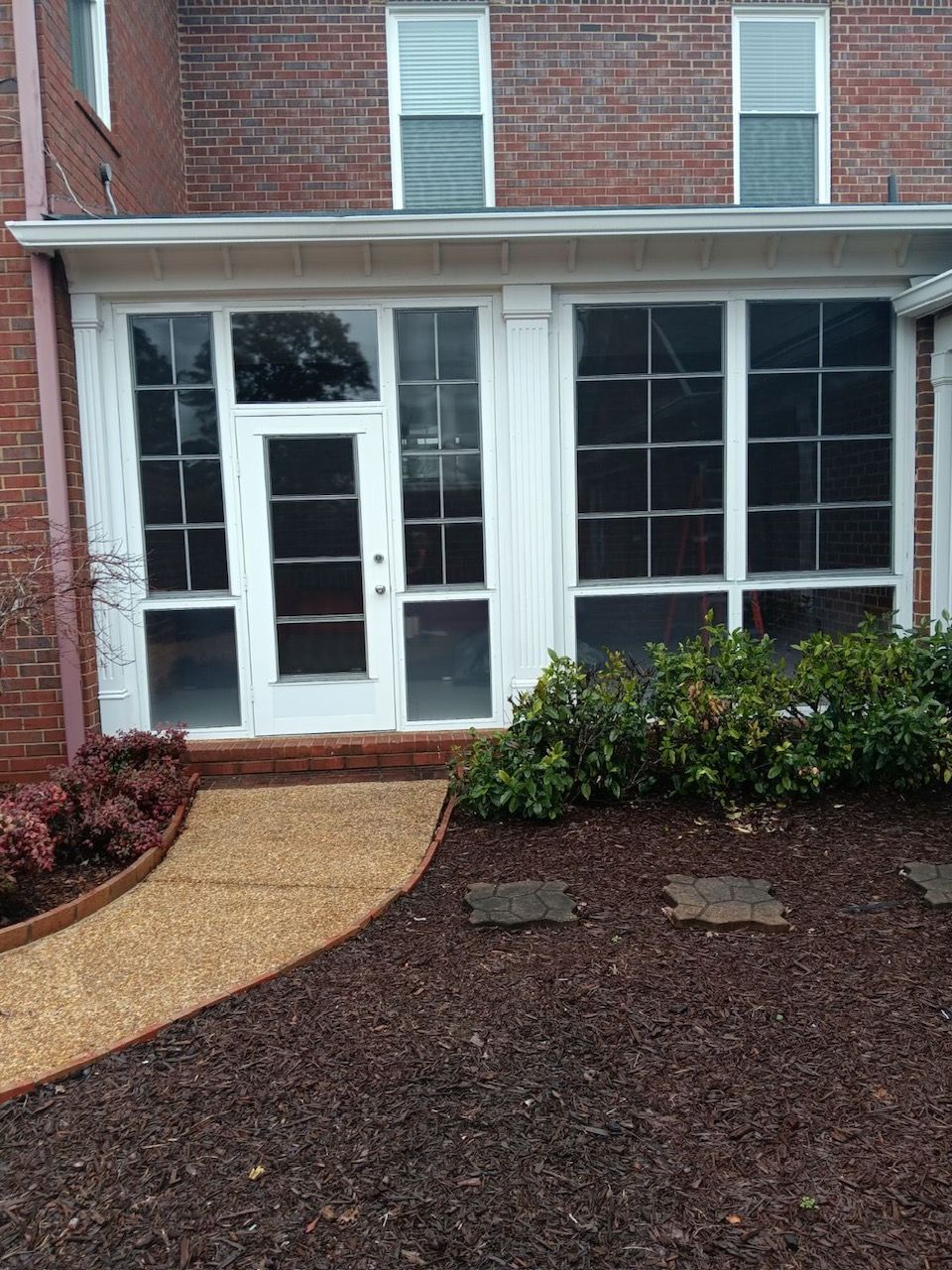 Brick apartment entrance with white door, large windows, and landscaped path with mulch and shrubs
