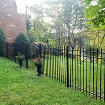 Black metal fence enclosing a grassy yard with brick building, trees, and potted flowers.