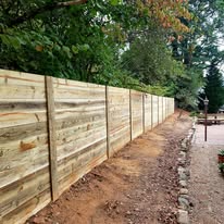 Wooden fence, light brown, borders a dirt path, green trees in background.