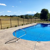 Swimming pool with blue water and metal handrails, next to a brown patio, fence, and a distant field.