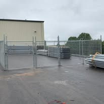Metal fencing with stacked metal items, near a building on a concrete surface under a cloudy sky.