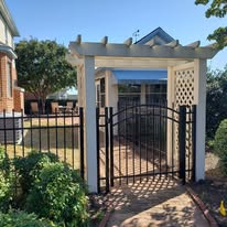 Arched black gate leading to a pergola entrance with lattice side panels and brick walkway.