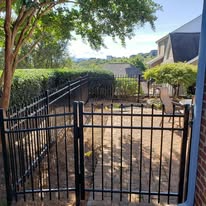 Black metal fence with gate, grassy area, trees, and house in the background on a sunny day.