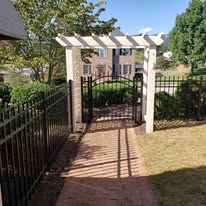 Black metal fence with arched gate and white pergola in front of a brick path.