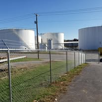 White storage tanks behind a chain-link fence, with power lines and a blue sky.