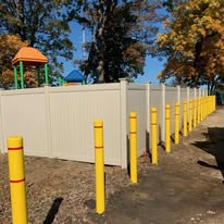 Yellow bollards line a walkway next to a tan fence, playground visible in the background.