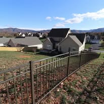 Brown metal fence in front of grassy hill, houses, and mountains on a sunny day.