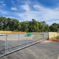 A chain-link fence with a gate surrounds a paved area, with trees and a blue sky in the background.