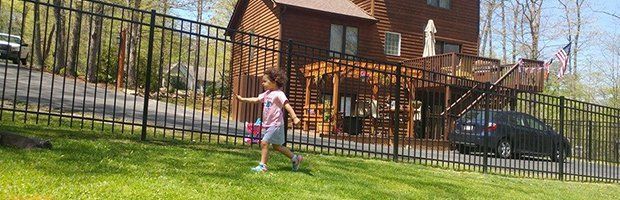 A child runs along a black fence in front of a house. The house is brown and has a deck. A car is parked nearby.