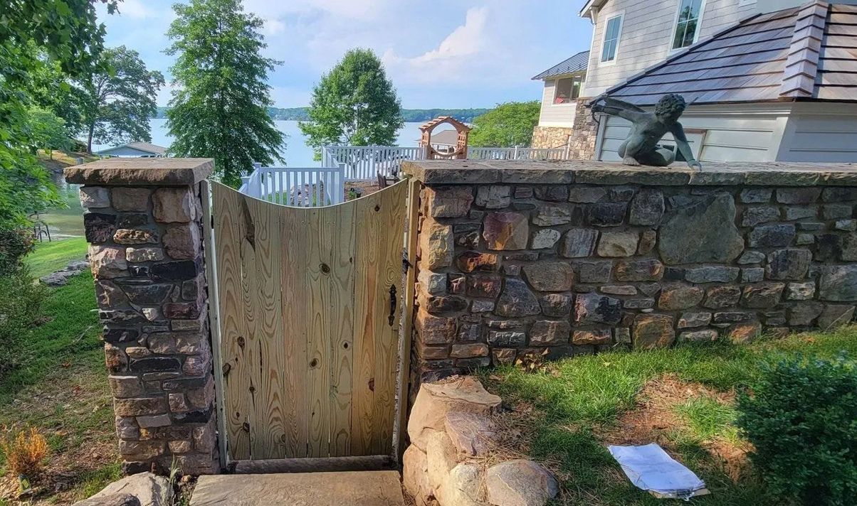 Stone wall with wooden gate leading to a lake. Trees and house in the background.