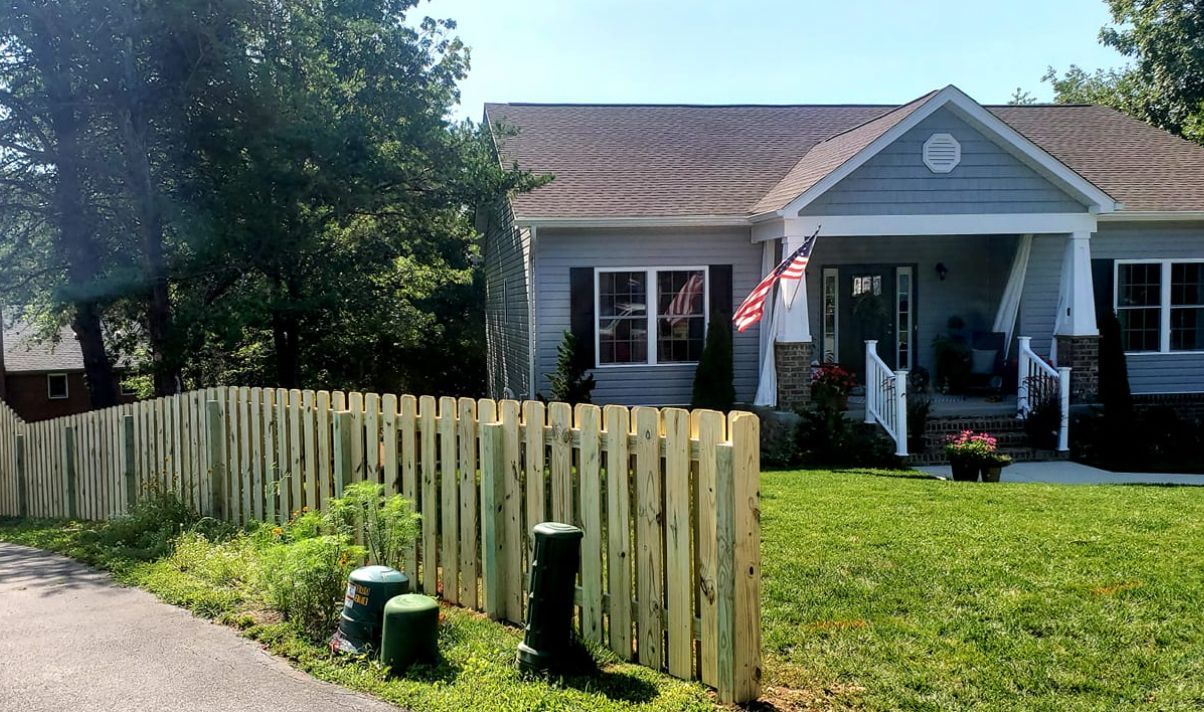 Cottage home with a wooden fence and American flag on the porch; green grass and clear sky.