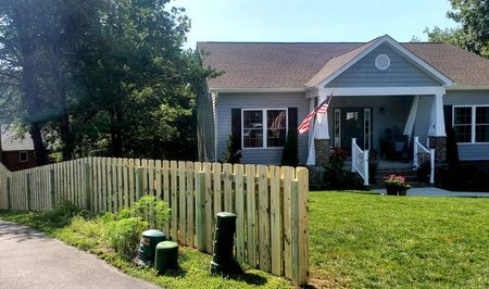 Cottage home with a wooden fence and American flag on the porch; green grass and clear sky.