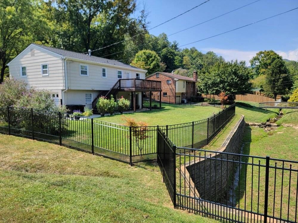 Two-story house with black metal fence surrounding a grassy yard next to a ditch. Blue sky, trees in background.