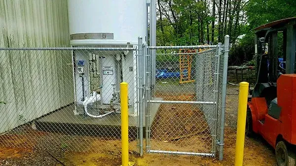 A fenced-in industrial area with a large white tank, yellow bollards, and an orange forklift.