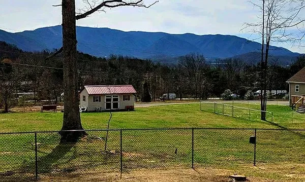 A small shed with a red roof sits in a grassy yard, with mountains in the background behind a chain-link fence.