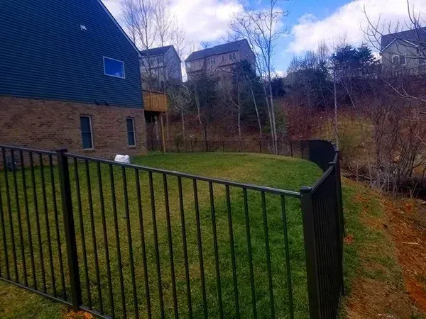 Black metal fence encloses a green lawn on a slight hillside. Houses and trees are in the background.