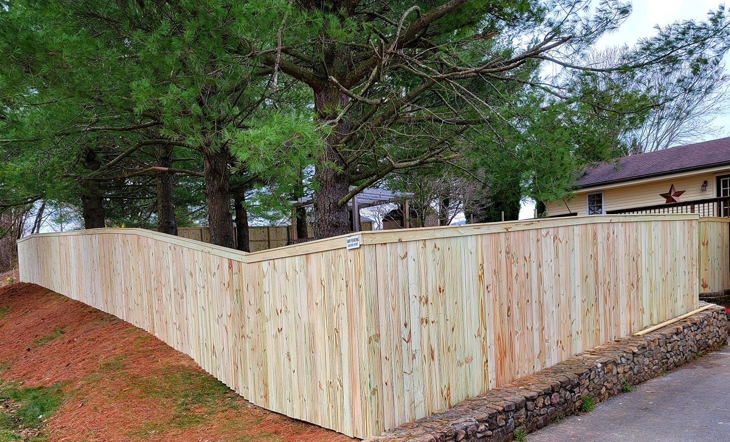 Wooden fence curves along a hillside with trees in the background and a house to the right.