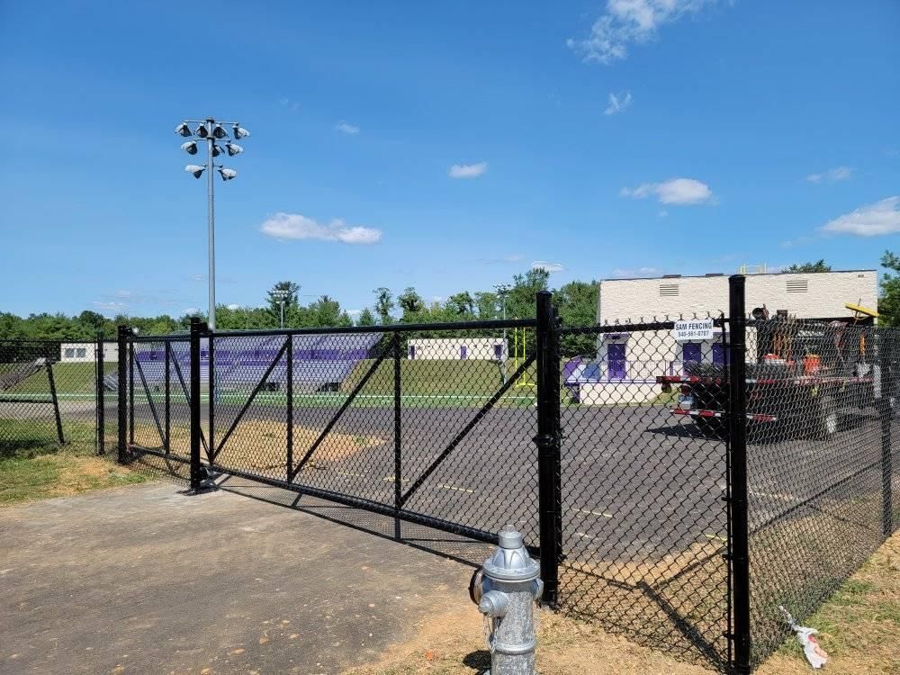 Black chain-link fence and gate enclosing a sports field; blue sky, white clouds, and light pole in the background.