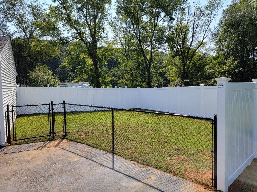 A backyard enclosed by a white vinyl fence and a black chain-link fence, with green grass and trees in the background.