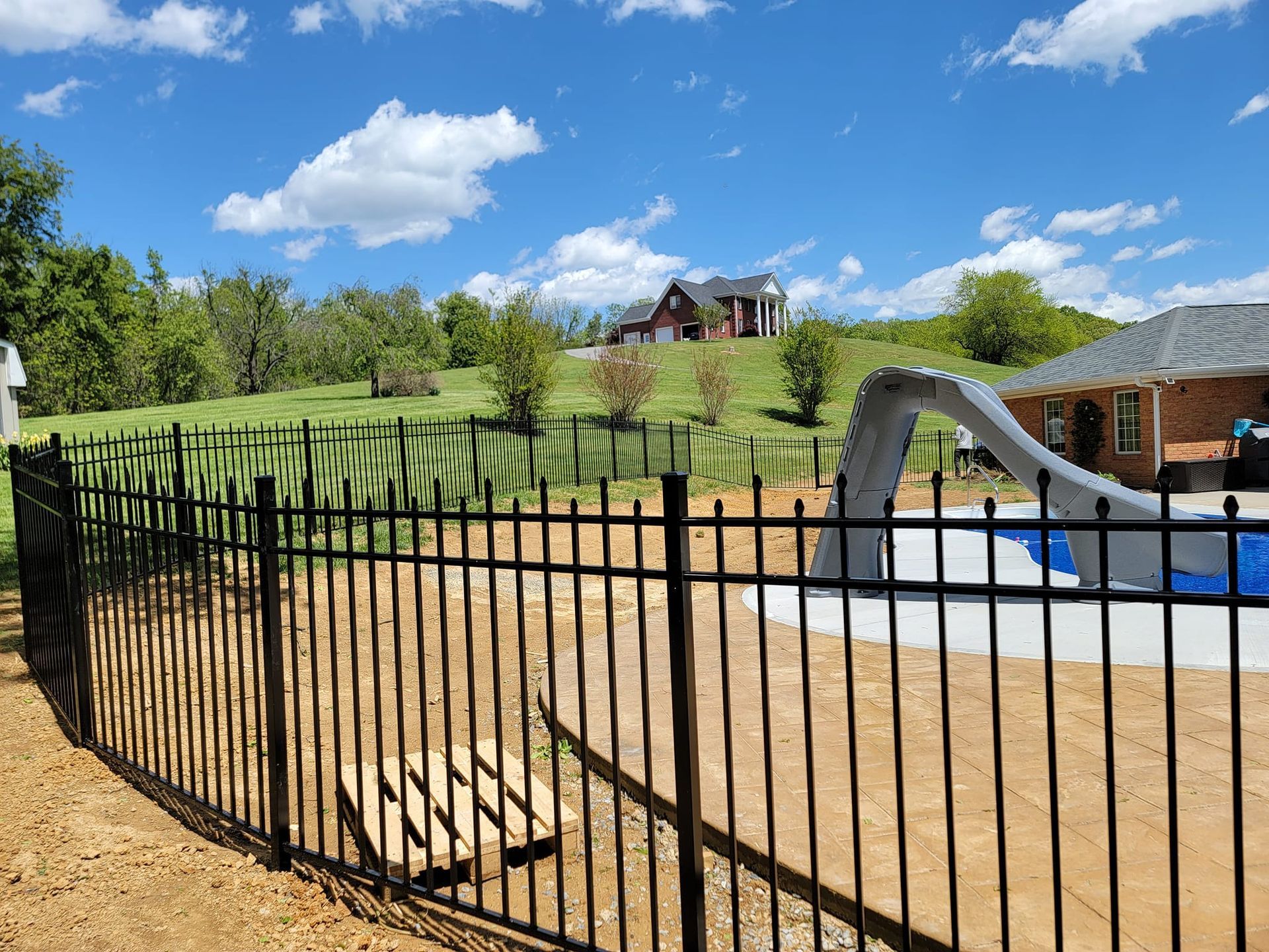 Black fence encloses a pool with a slide, hillside in the background, under a blue sky with clouds.
