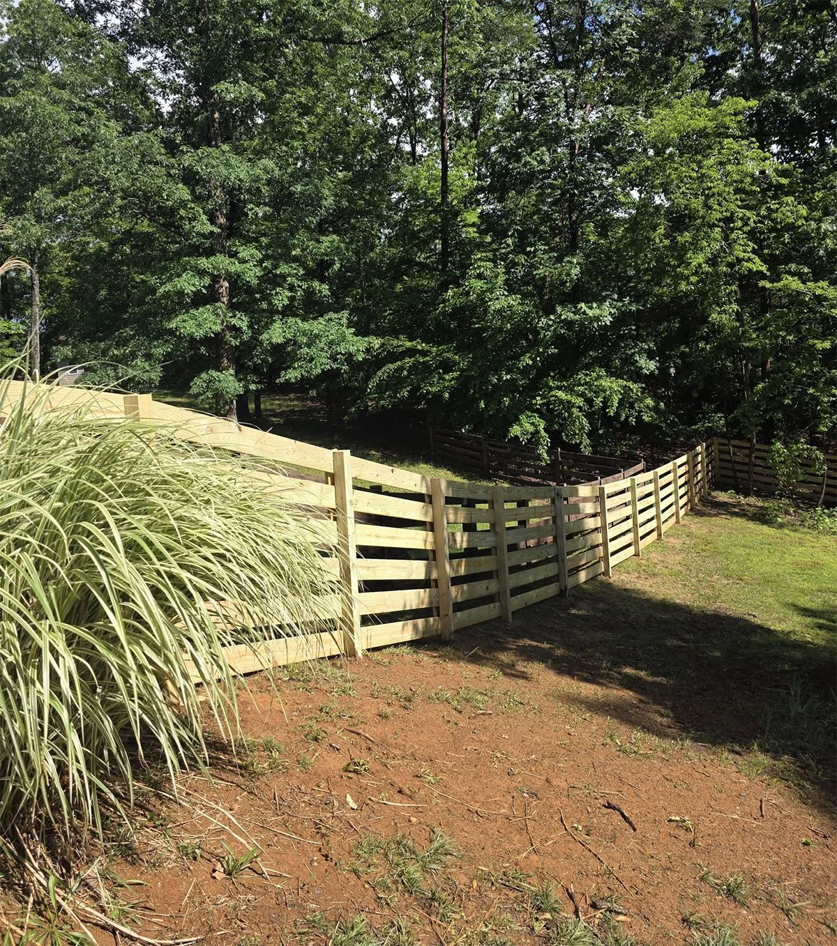 Wooden horizontal-slat fence on a hillside, separating a yard from a wooded area. Green foliage in background.