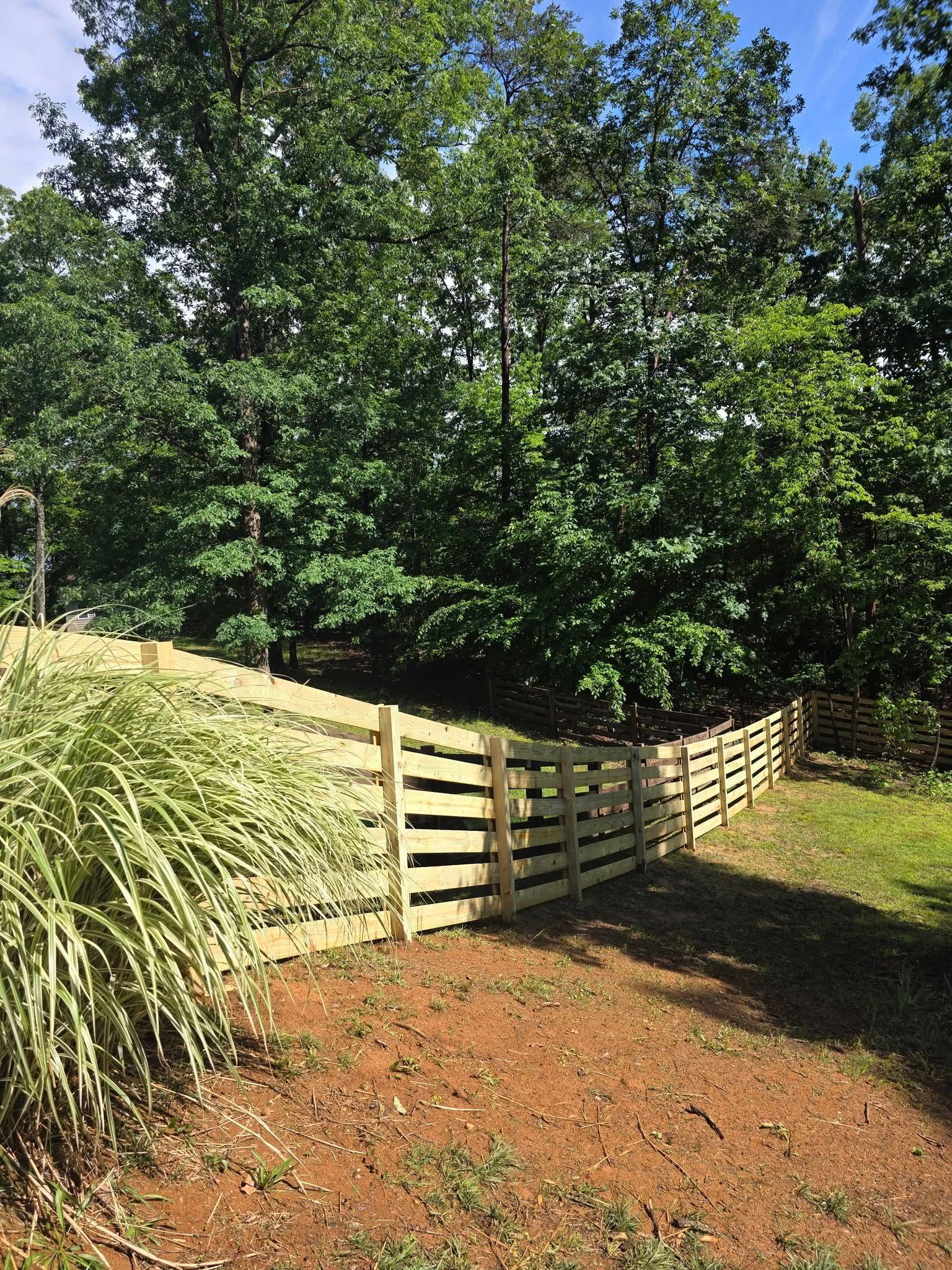 Wooden fence on a slight incline, bordering a yard. Tall trees are in the background. Sunny day.