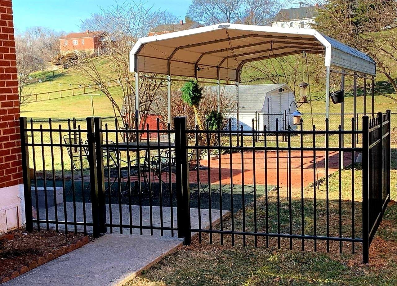 Black metal fence encloses a small patio with a carport in a grassy yard.