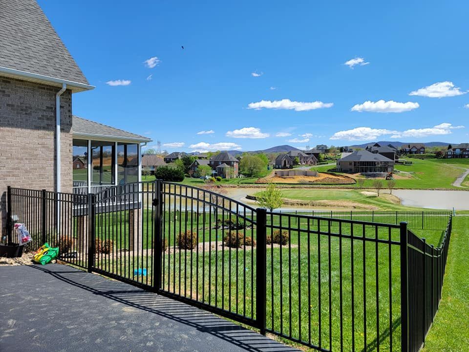 Black metal fence surrounding a grassy yard with a view of houses, a pond, and a blue sky.