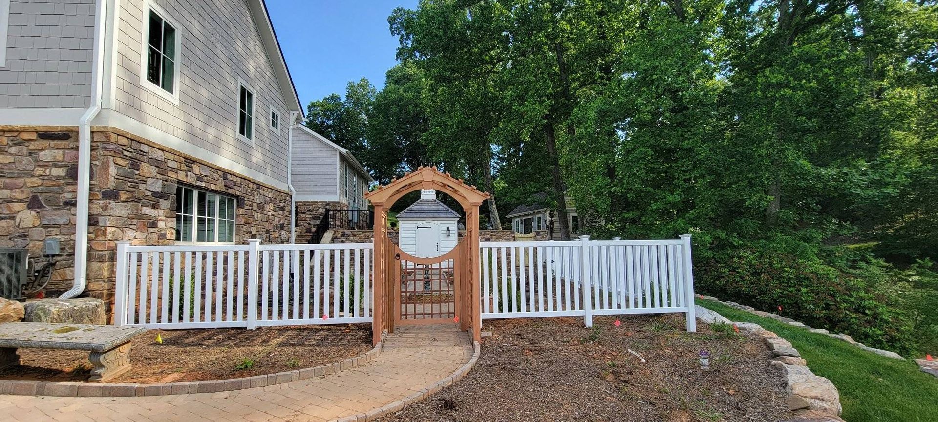A wooden arched gate in a white picket fence leads to a path in front of a building with stone siding.