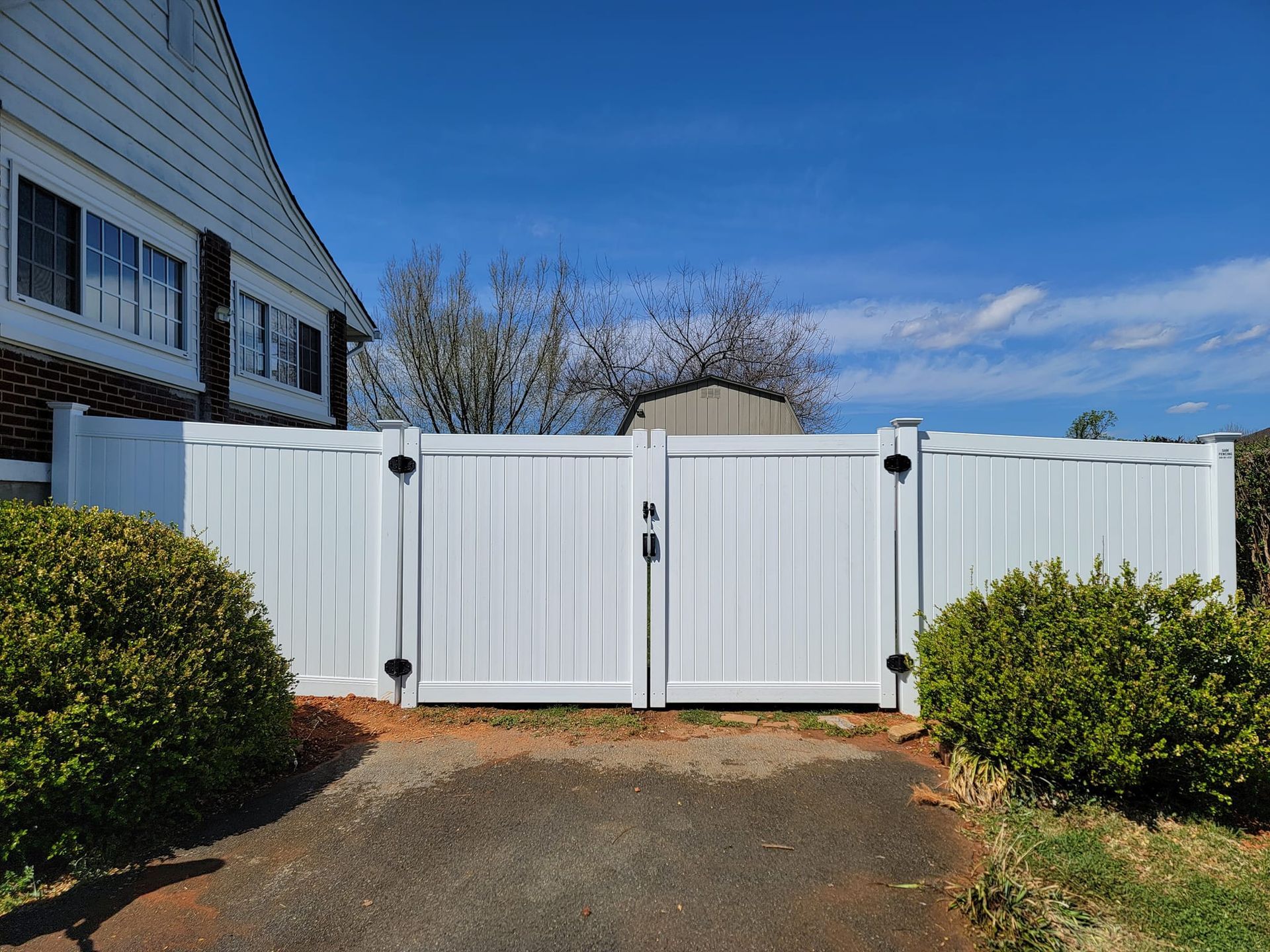 White double-door gate in a white fence, in front of a house, set on a sunny day.