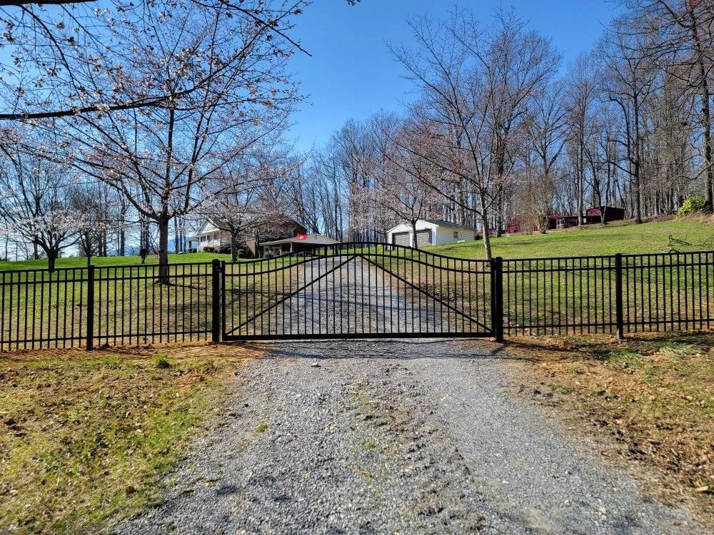 Black metal gate and fence on a gravel driveway leading to a house, with trees and blue sky.