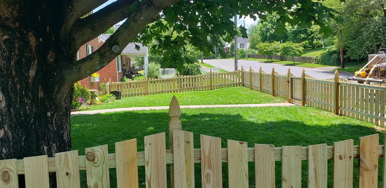 A wooden picket fence surrounds a grassy yard near a street, with a large tree on the left.