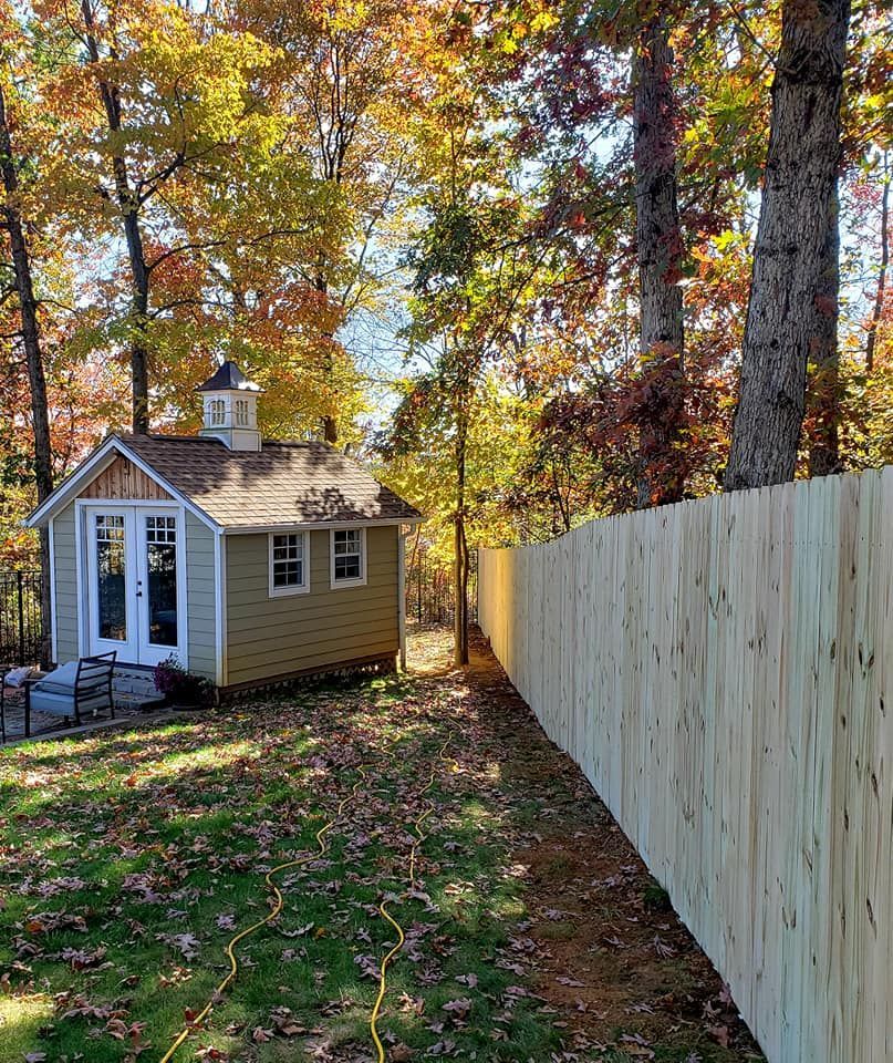 Small tan shed with cupola, next to a wooden fence, in a yard with fall leaves.