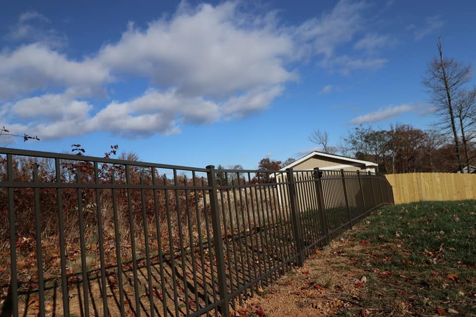 Black metal fence in front of a yard with a wooden fence and a house in the background under a blue sky with clouds.