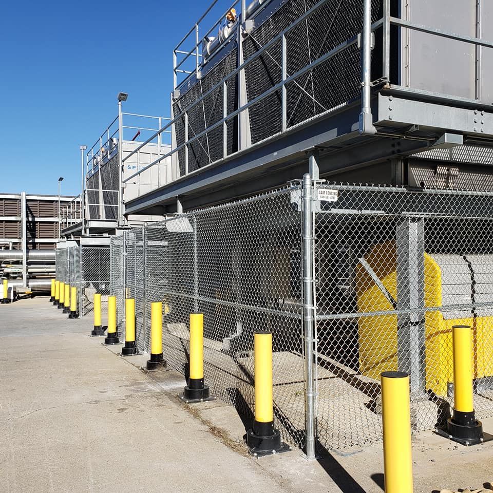 Chain-link fence with yellow bollards in front of industrial cooling towers on a sunny day.