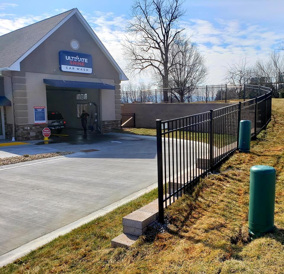 Car wash exterior with black fence, retaining wall, and green grass. Blue sky.
