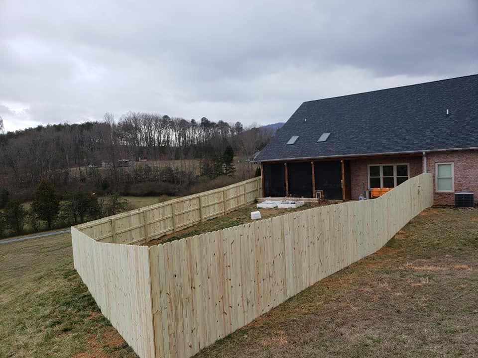 Wooden fence encloses a backyard with a house, on a grassy hillside under a cloudy sky.